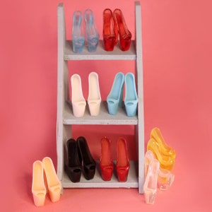 Puede incluir: Un conjunto de diez pares de zapatos de tacón alto en varios colores, incluyendo rojo, azul, blanco, negro y transparente, exhibidos en una estantería de madera.