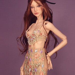 May include: A doll wearing a beige lace top and skirt with floral embroidery in shades of pink, blue, and green. The doll has long, wavy red hair and is posed with one hand on her hip.