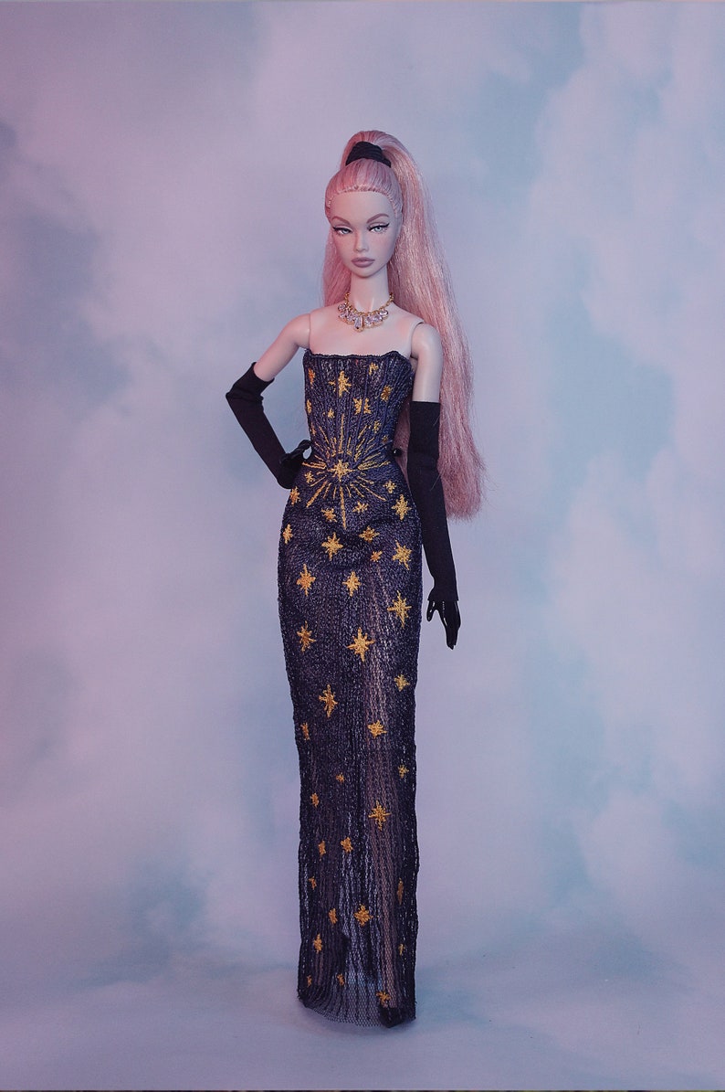May include: A doll wearing a black gown with gold star embroidery and black gloves. The doll has pink hair and is wearing a necklace.