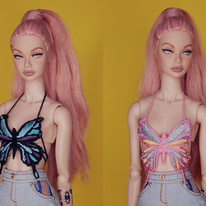 May include: Two dolls with pink hair wearing halter tops with butterfly embroidery. One top is blue and green, the other is pink and yellow. Both dolls are wearing denim shorts.