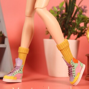 May include: A pair of mustard yellow socks with a doll wearing colorful high-top sneakers. The doll's legs are crossed and the socks are pulled up to the knees. The background is a pink wall with a potted plant.