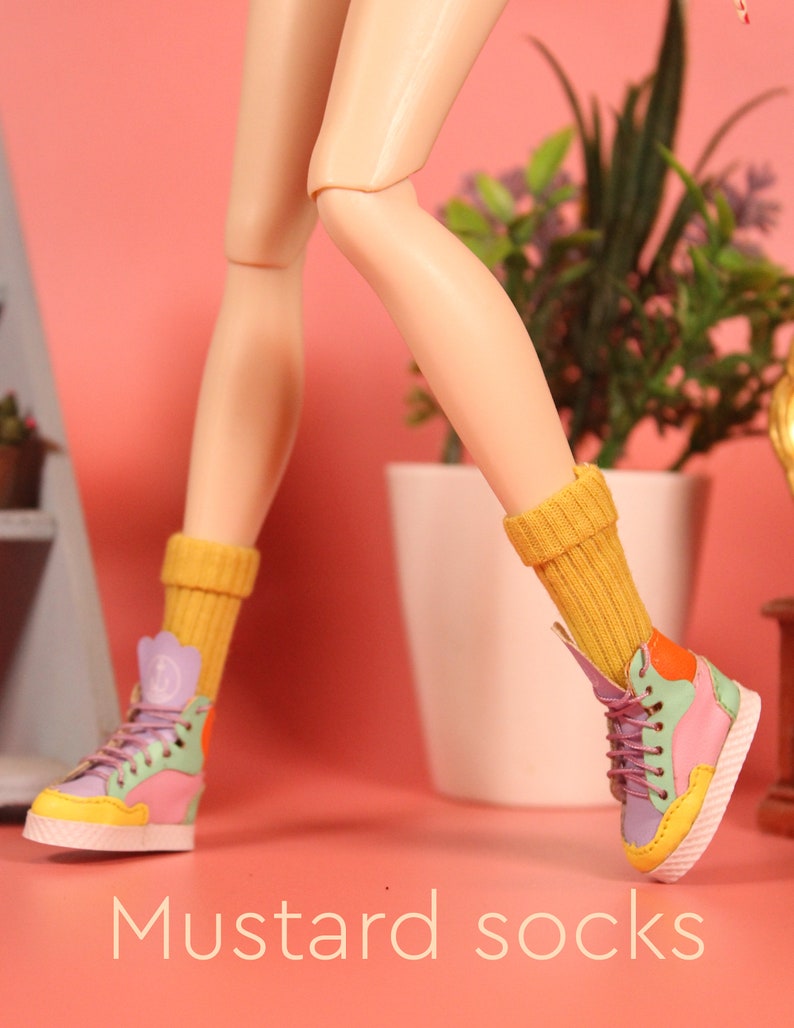 May include: A pair of mustard yellow socks with a doll wearing colorful high-top sneakers. The doll's legs are crossed and the socks are pulled up to the knees. The background is a pink wall with a potted plant.
