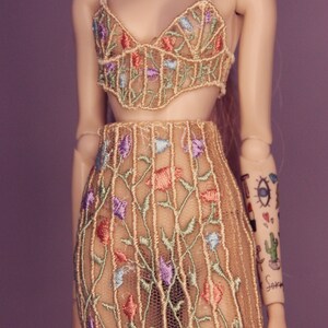 May include: A beige mesh top and skirt set with floral embroidery in shades of blue, green, and pink. The top has a sweetheart neckline and thin straps. The skirt is a mini skirt with a high waist.