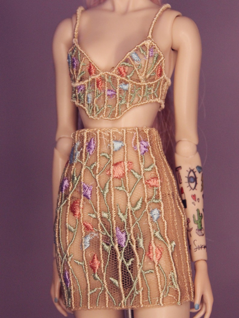May include: A beige mesh top and skirt set with floral embroidery in shades of blue, green, and pink. The top has a sweetheart neckline and thin straps. The skirt is a mini skirt with a high waist.