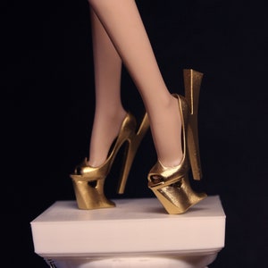 May include: A pair of gold high-heeled shoes with a unique platform design. The shoes are on a white pedestal.