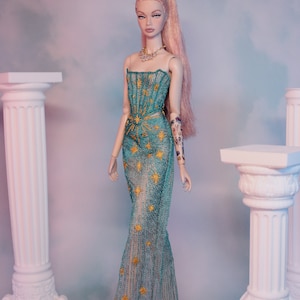 May include: A fashion doll with long pink hair styled in a ponytail, wearing a teal gown with gold star embellishments and a gold necklace. The doll is standing in front of two white columns.