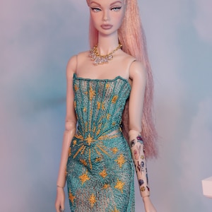 May include: A doll wearing a teal and gold star-patterned dress with a sheer overlay. The doll has pink hair pulled back in a ponytail and a diamond necklace. The doll's right arm has a tattoo with various designs, including a peace sign, a heart, and the word "love".