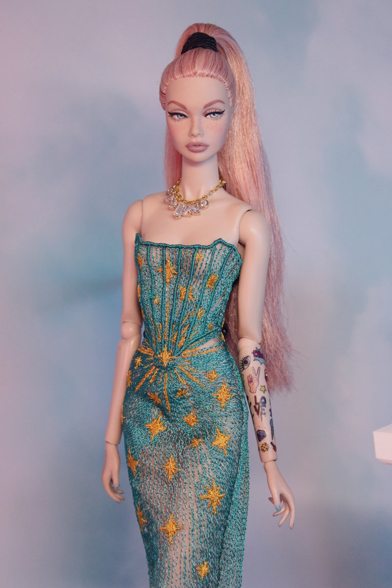 May include: A doll wearing a teal and gold star-patterned dress with a sheer overlay. The doll has pink hair pulled back in a ponytail and a diamond necklace. The doll's right arm has a tattoo with various designs, including a peace sign, a heart, and the word "love".