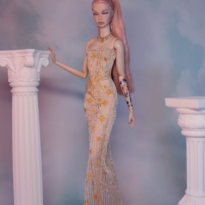 May include: A doll wearing a gold and white beaded gown with a star pattern. The doll has pink hair and is standing in front of two white columns.