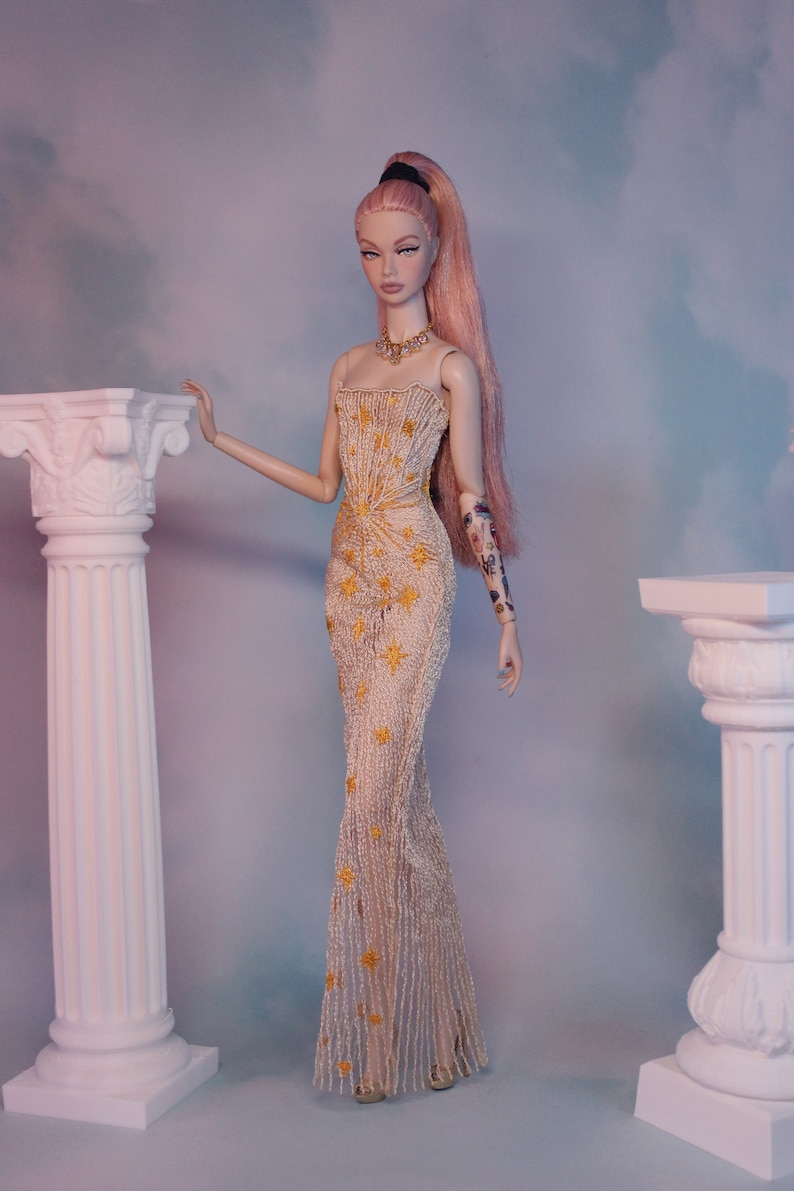 May include: A doll wearing a gold and white beaded gown with a star pattern. The doll has pink hair and is standing in front of two white columns.
