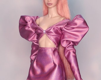 May include: A fashion doll with pink hair and makeup, wearing a metallic pink mini dress with a cut-out and large puffed sleeves. The doll is posed against a light blue and white background.