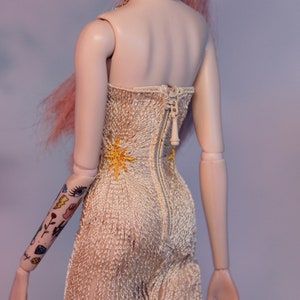 May include: A doll wearing a white, crocheted, strapless dress with a gold star design. The dress has a zipper down the back. The doll has pink hair and a gold chain necklace.