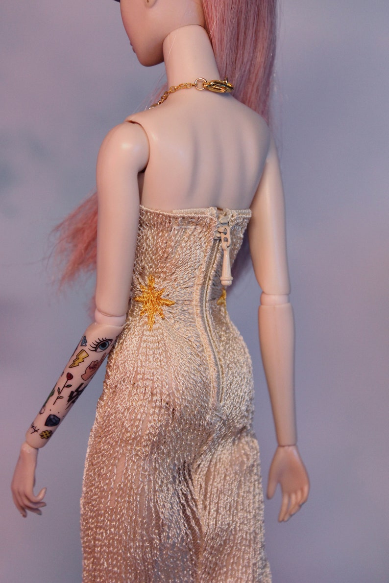 May include: A doll wearing a white, crocheted, strapless dress with a gold star design. The dress has a zipper down the back. The doll has pink hair and a gold chain necklace.