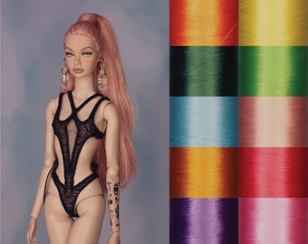 May include: A fashion doll with pink hair wearing a black lace bodysuit with cutouts. The doll has large earrings and a tattoo on her arm. The background is a blurred image of colorful spools of thread.