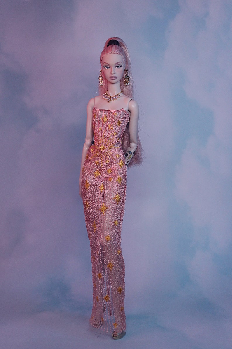 May include: A pink and gold sequined gown with a star pattern. The gown has a strapless bodice and a flowing skirt. The doll is wearing gold earrings and a gold necklace.