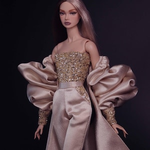May include: A fashion doll with long two-toned hair wearing a champagne-colored jumpsuit featuring a sequined bodice and waist accents. The outfit includes a matching long-sleeved coat with voluminous, gathered sleeves and sequined cuffs.