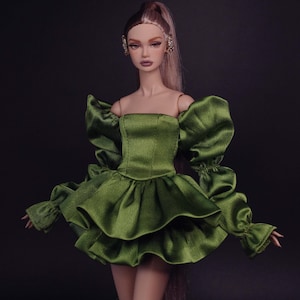 May include: A fashion doll with long light brown hair styled in a high ponytail, wearing a short olive green satin dress. The dress features a structured bodice, voluminous puffed sleeves with gathered cuffs, and a layered, ruffled skirt.