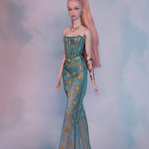May include: A fashion doll with long pink hair, wearing a teal blue gown with gold star embroidery. The doll has a delicate gold necklace and a tattoo on her arm.