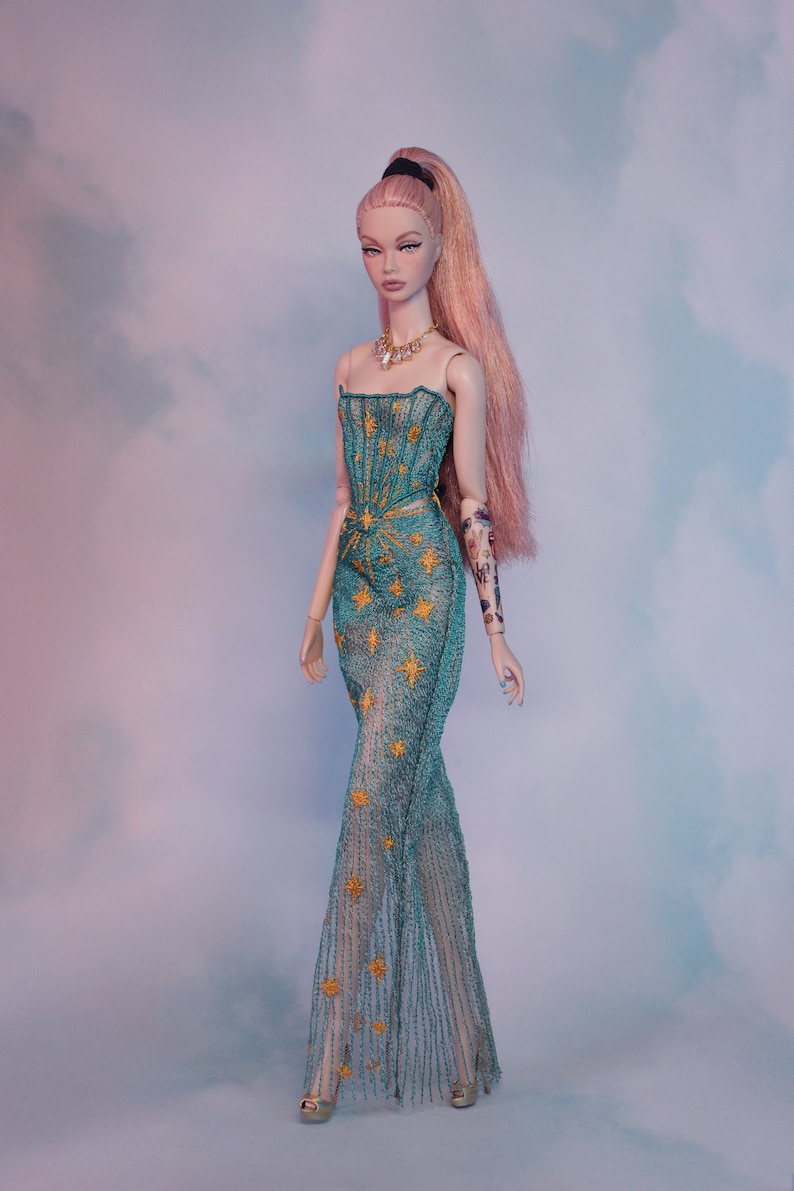 May include: A fashion doll with long pink hair, wearing a teal blue gown with gold star embroidery. The doll has a delicate gold necklace and a tattoo on her arm.