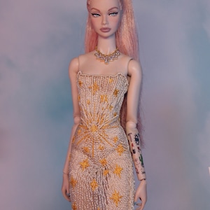 May include: A doll with pink hair styled in a high ponytail, wearing a white dress with gold star embroidery and a diamond necklace.
