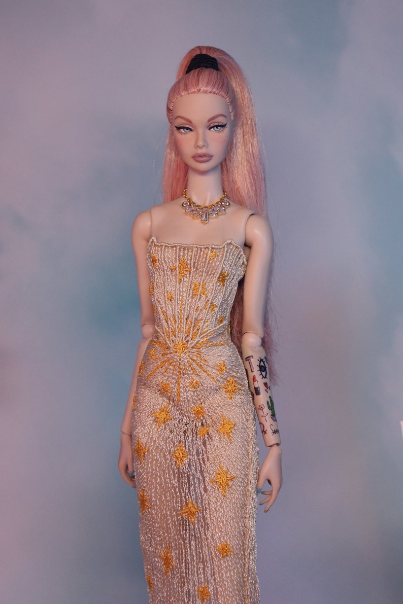 May include: A doll with pink hair styled in a high ponytail, wearing a white dress with gold star embroidery and a diamond necklace.