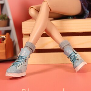 May include: A pair of blue socks with white anchors on them are worn by a doll with light skin. The doll is wearing blue denim shorts and is sitting on a wooden crate.
