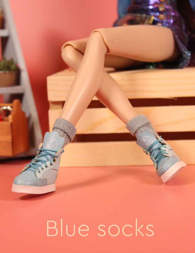 May include: A pair of blue socks with white anchors on them are worn by a doll with light skin. The doll is wearing blue denim shorts and is sitting on a wooden crate.