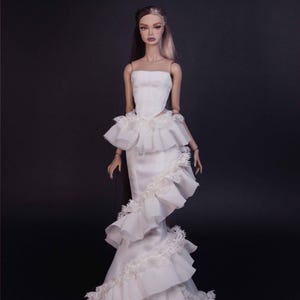 May include: A fashion doll with two-toned hair wearing a strapless white corset bodice and a floor-length skirt featuring cascading ruffled layers and delicate lace trim. The doll has a neutral expression and articulated limbs, standing against a dark background.