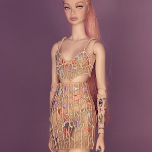 May include: A doll wearing a beige, floral embroidered dress with a sheer fabric. The doll has pink hair and a tattoo on her arm.