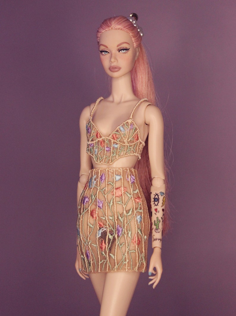 May include: A doll wearing a beige, floral embroidered dress with a sheer fabric. The doll has pink hair and a tattoo on her arm.