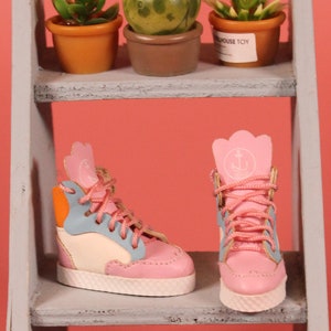 May include: Two pairs of pink high-top sneakers with white soles. One pair has blue, orange, and white accents. The other pair has a white anchor logo on the side. The sneakers are on a wooden shelf with small potted plants.