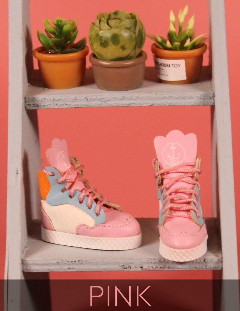 May include: Two pairs of pink high-top sneakers with white soles. One pair has blue, orange, and white accents. The other pair has a white anchor logo on the side. The sneakers are on a wooden shelf with small potted plants.
