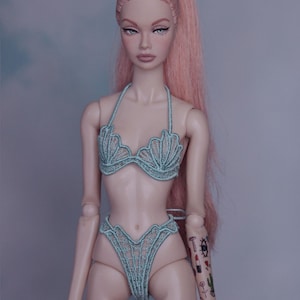May include: A doll with pink hair in a ponytail wearing a light blue bikini with a seashell design. The doll has a tattoo on its arm with a cactus, lipstick, and an eye.