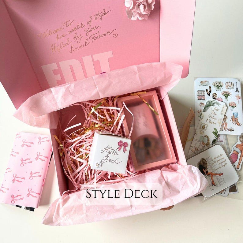May include: A pink gift box with a white tissue paper lining. The box contains a small pink box with the words "Style Deck" written on it. The box is surrounded by pink and gold shredded paper. The box is sitting on a white surface.