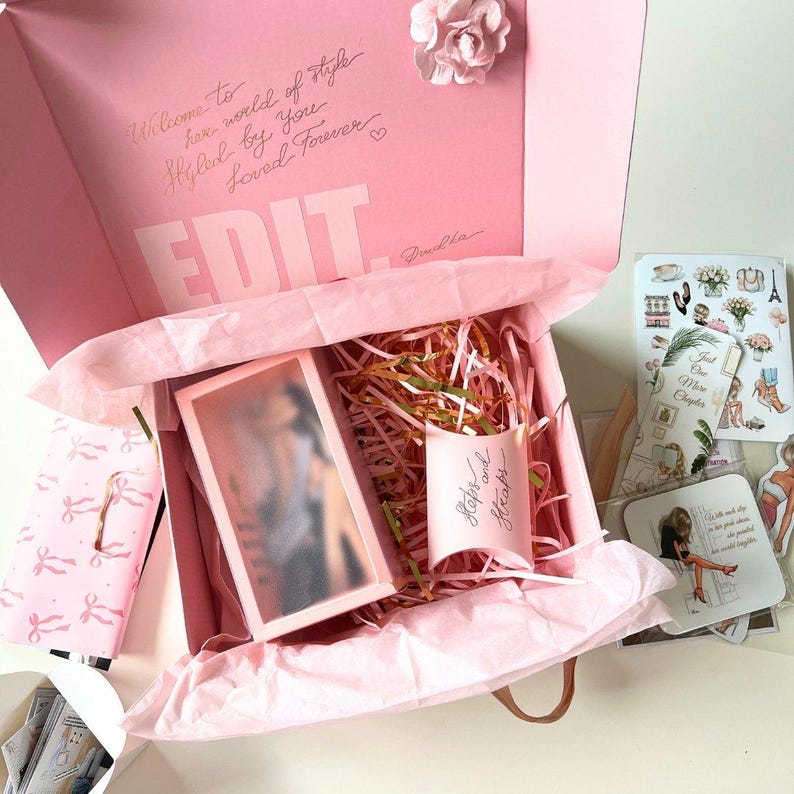 May include: A pink gift box with a white box inside. The gift box is filled with pink tissue paper and gold and white shredded paper. The white box has a clear lid and a picture of a person inside. The gift box has the words "Welcome to the world of style" and "Styled by you" written on the top. The gift box also has the word "EDIT" written on the front.