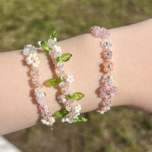 May include: Three beaded bracelets with floral and leaf designs. The bracelets feature pink, white, and clear beads, with green leaf accents. The bracelets are worn on a wrist, showcasing the delicate craftsmanship and the variety of floral patterns.