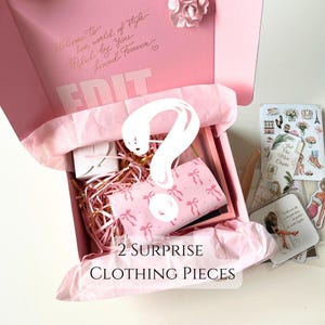 May include: A pink box with the words "Welcome to her world of style Shiled by You Loved Forever EDIT" on the lid. The box is open and filled with pink tissue paper. There is a question mark drawn on the tissue paper. The box contains two surprise clothing pieces.