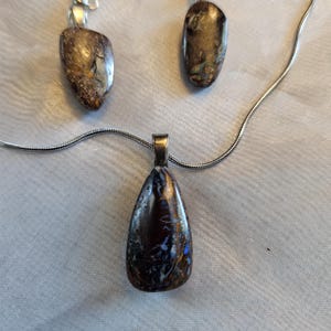May include: A jewelry set featuring a pendant necklace and matching earrings. The pendant and earrings have a dark, iridescent stone with brown, blue, and gold tones. The necklace has a silver chain, and the earrings have silver hooks.