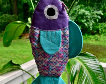 May include: A whimsical, handmade fish-shaped bag. The bag features a purple head with a large eye, teal fins, and a body adorned with a scale pattern in shades of pink, blue, and purple. It has a white strap for carrying.