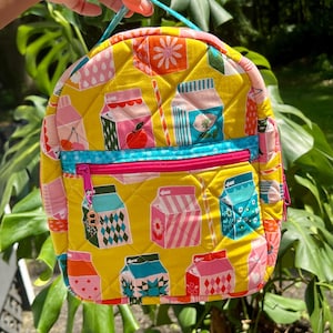 May include: A quilted yellow backpack with a repeating pattern of milk carton illustrations in various colors. The backpack has a blue zipper pocket, a pink zipper, and a blue handle. The design includes a variety of colors, including pink, blue, and orange.