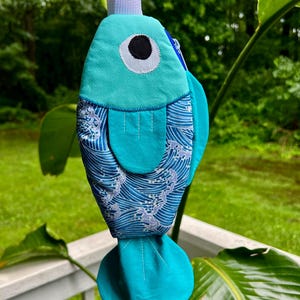 Puede incluir: Una bolsa de tela con forma de pez turquesa con una correa blanca. La bolsa presenta un ojo grande con una pupila negra e iris blanco. El cuerpo tiene un patrón de olas en azul y blanco. Las aletas y la cola son turquesas.