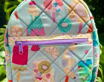 May include: A small, quilted backpack with a light blue background and a colorful pattern of ice pops, strawberries, and kiwi slices. The backpack has a purple zipper and a small handle at the top. The bag is approximately 10 inches tall.