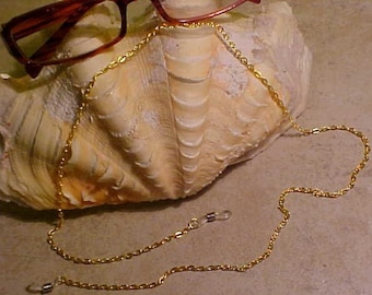 May include: A gold chain eyeglass retainer with clear plastic end pieces. The chain is lying on a seashell.