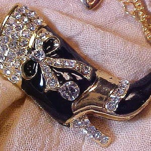May include: A gold-tone metal pendant shaped like a cowboy boot, featuring a glossy black enamel finish, clear rhinestone accents on the cuff, heel, and a decorative bow detail on the front, suspended from a matching gold-tone link chain necklace.