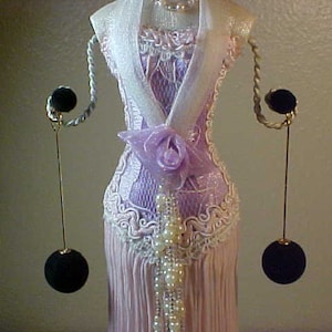 May include: A white dress form with a pink and white lace bodice and a white satin sash. The dress form is wearing a pair of black earrings with dangling balls.