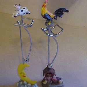 May include: Two silver metal stands with decorative figurines. The stand on the left has a black and white cow figurine and a yellow crescent moon. The stand on the right has a yellow rooster figurine and a red barn figurine.