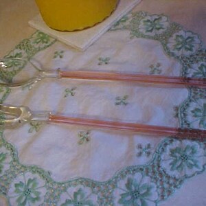 May include: A set of two glass salad servers with pink handles. The servers are on a white tablecloth with green embroidered flowers.