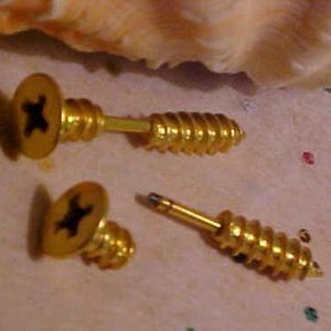 May include: Three gold screw-shaped earrings. The earrings are designed to look like screws with a flat head and a threaded shaft. The heads of the earrings are black.
