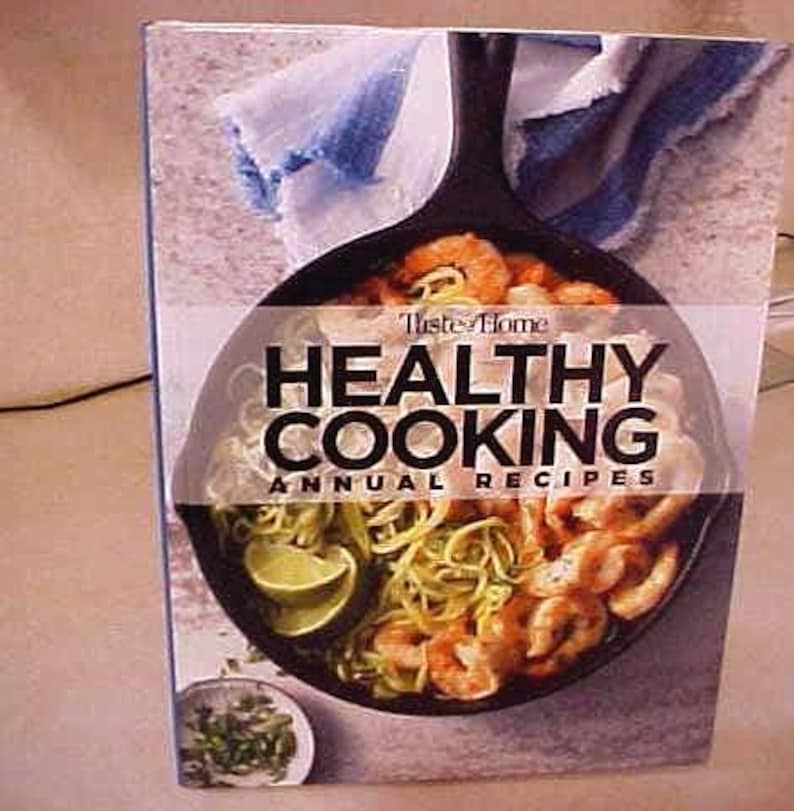 K&ouml;nnte beinhalten: Ein Kochbuch mit dem Titel "Taste of Home Healthy Cooking Annual Recipes" zeigt eine Pfanne mit Garnelen, Nudeln und Limette. Das Cover hat ein Foto des Gerichts mit dem Titel dar&uuml;ber. Eine kleine Sch&uuml;ssel mit Kr&auml;utern ist unten sichtbar.