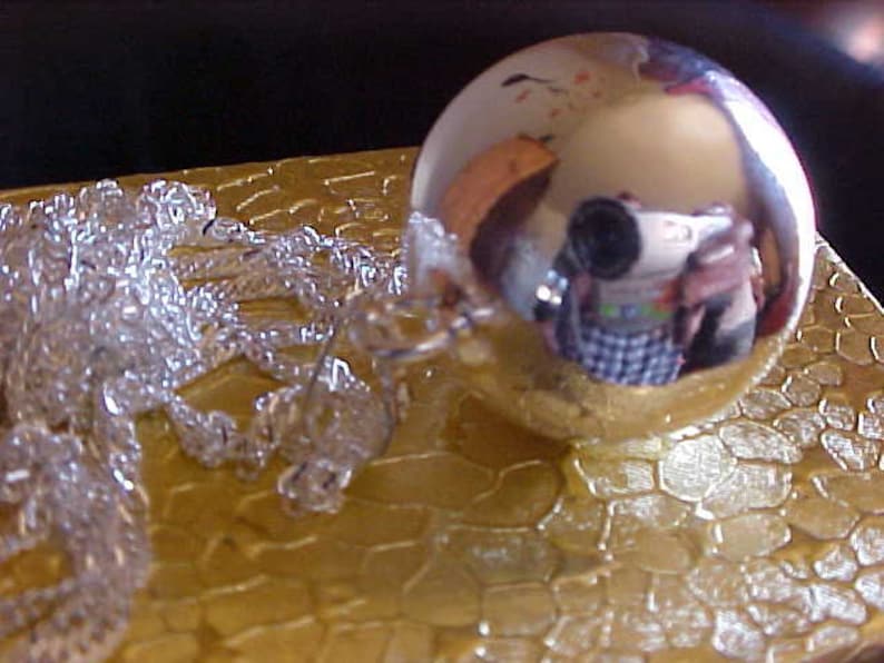 May include: A silver-colored, round, decorative ornament with a reflective surface. The ornament is sitting on a gold-colored, textured surface.